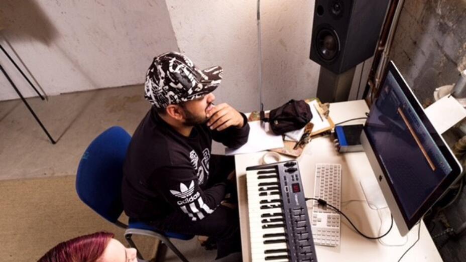A young man and a woman are at a desk in a recording studio. They are in front of a keyboard and a computer monitor