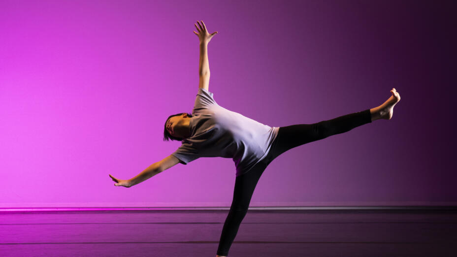 A dancer is in a bright pink studio. They have one foot on the ground but the rest of their limbs outstretched, mid-dance