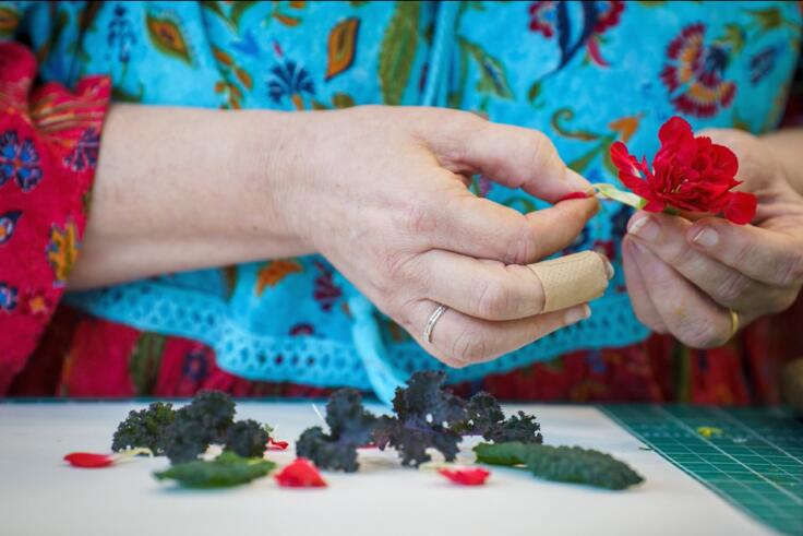 Close up of a woman in a turquoise floral top with red sleeves doing delicate craft work and holding a red fabric flower.