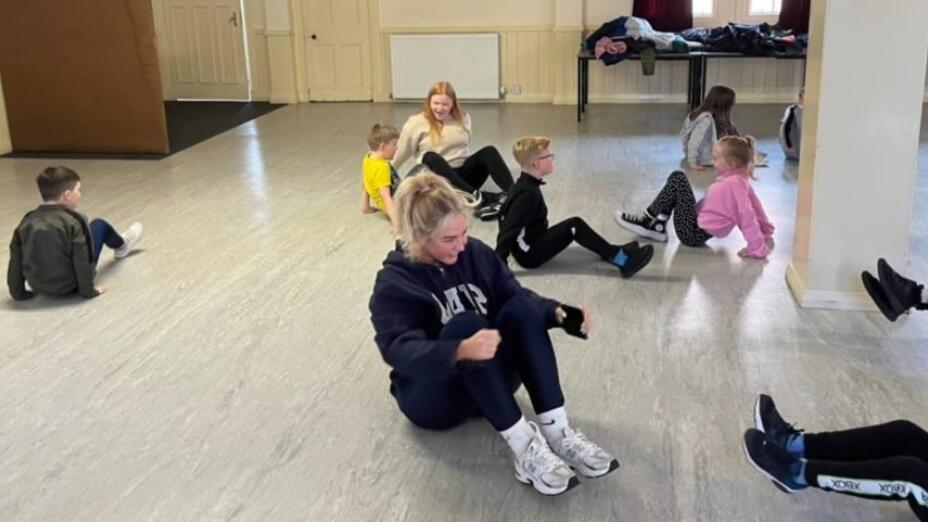 Two arts practitioners and nine pupils sit on the floor in bright room and explore learning through dance at St Mary’s RC Primary School.