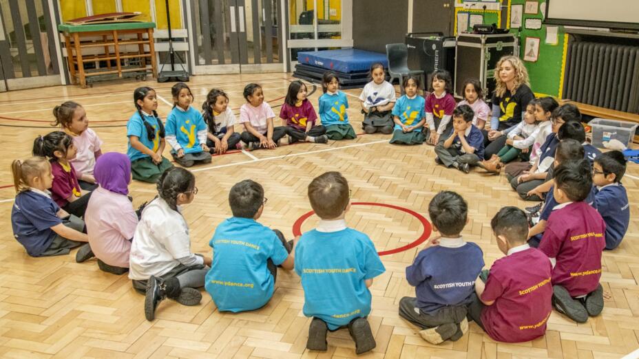 A group of children sit in a circle in a school gym during a workshop with an adult dance artist.